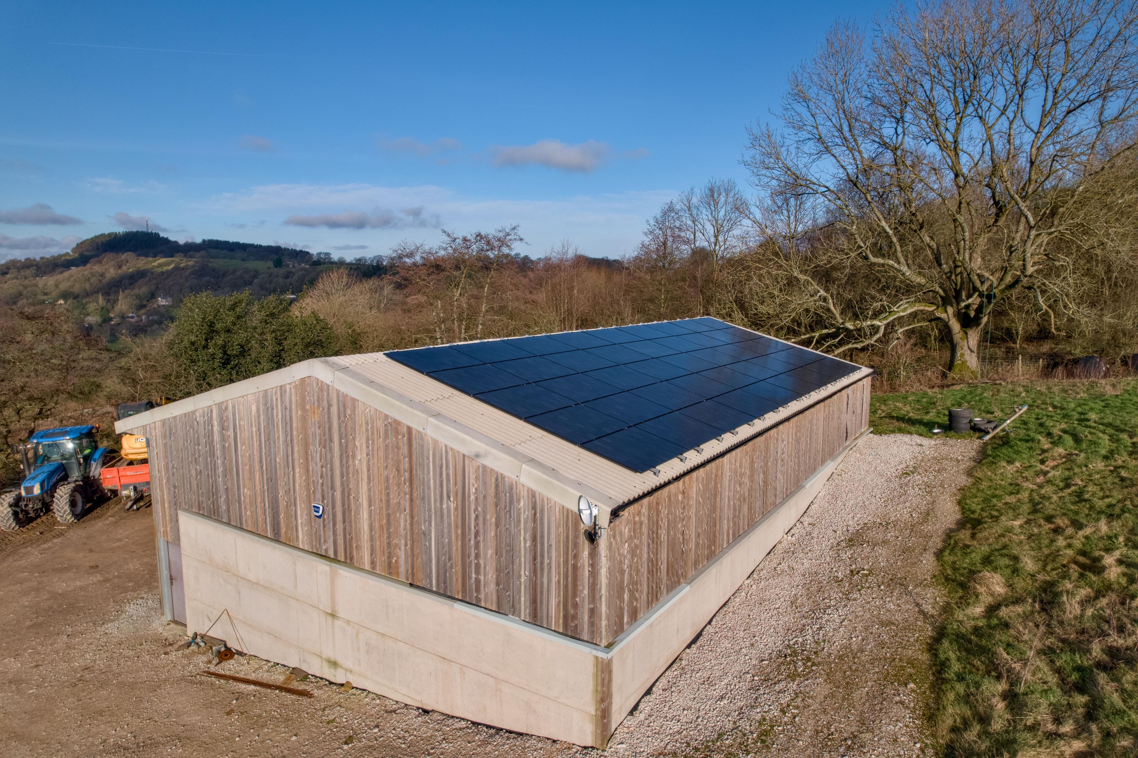 Black solar panels covering one roof slope of a timber-clad agricultural building in hilly countryside.