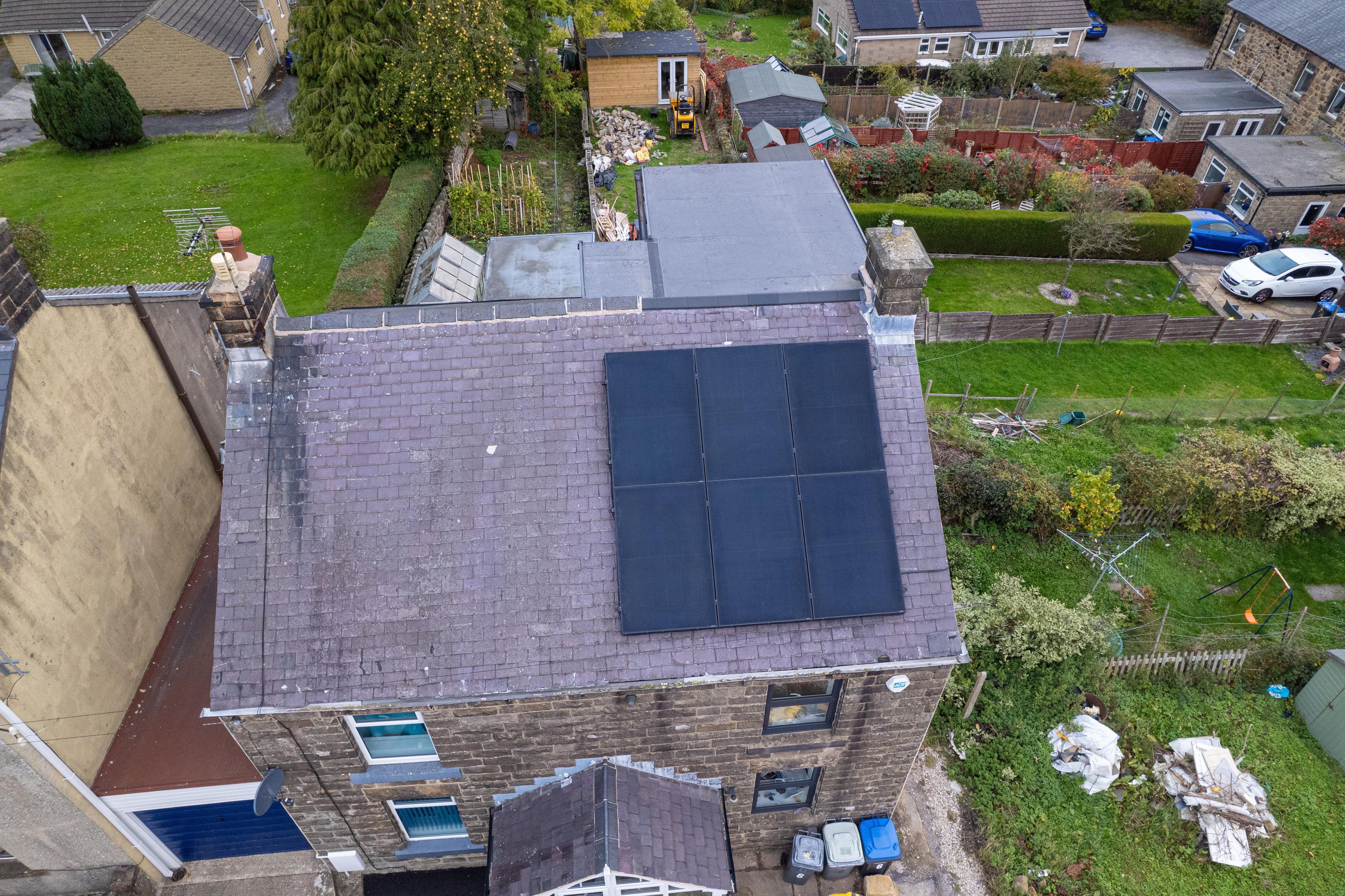 Aerial view of six black solar panels on the slate roof of a stone house with gardens behind.