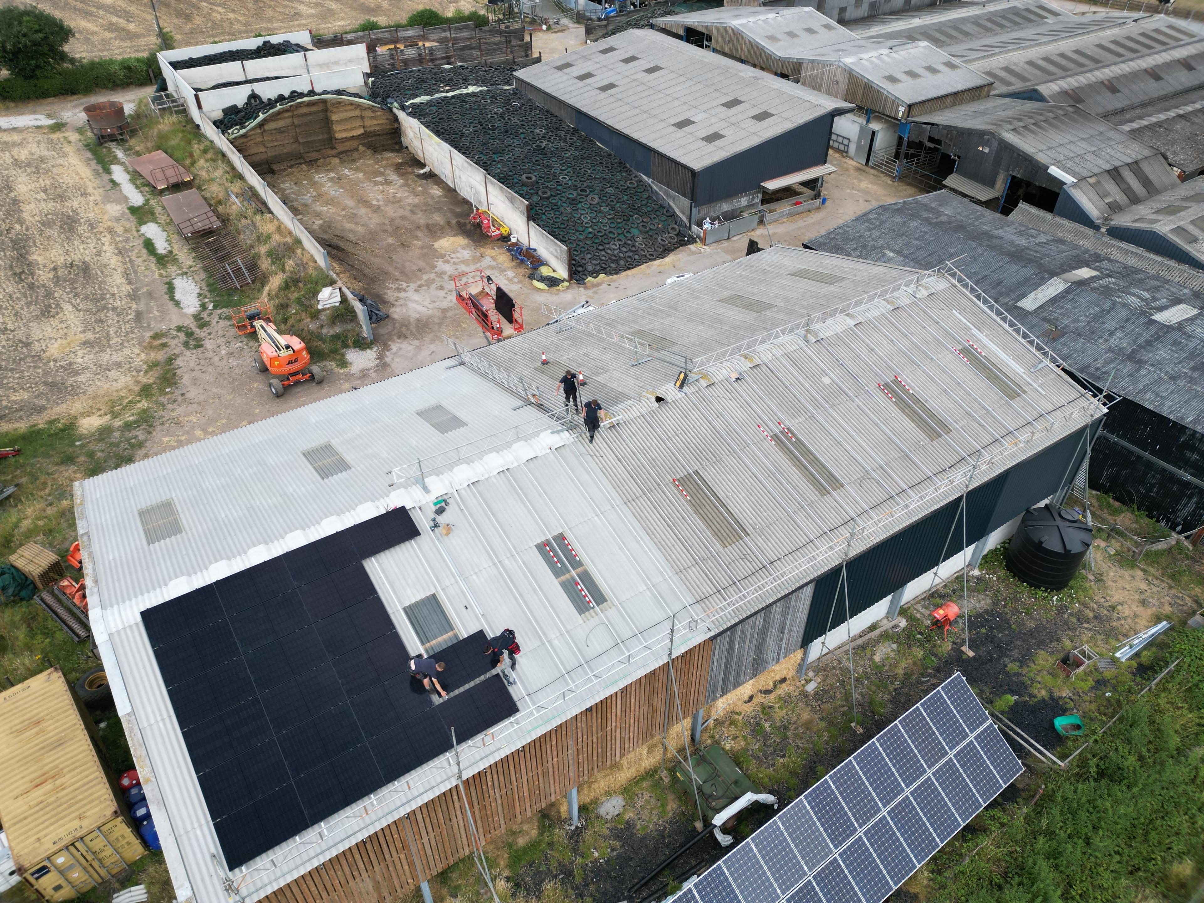Installers fitting black solar panels on a large farm building roof, with a ground-mounted array below.