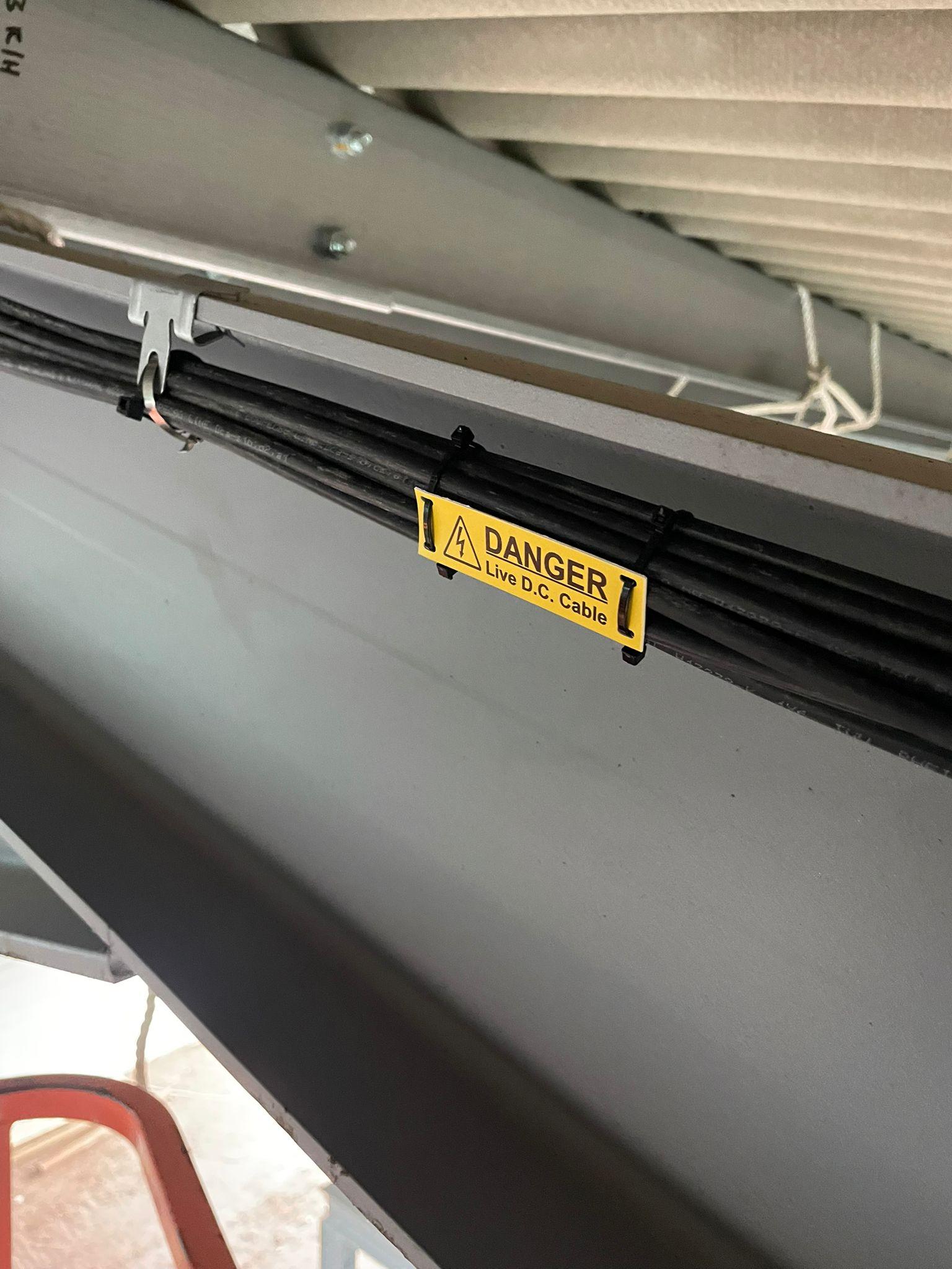 Bundle of black cables clipped under a steel beam with a yellow live DC cable danger label.