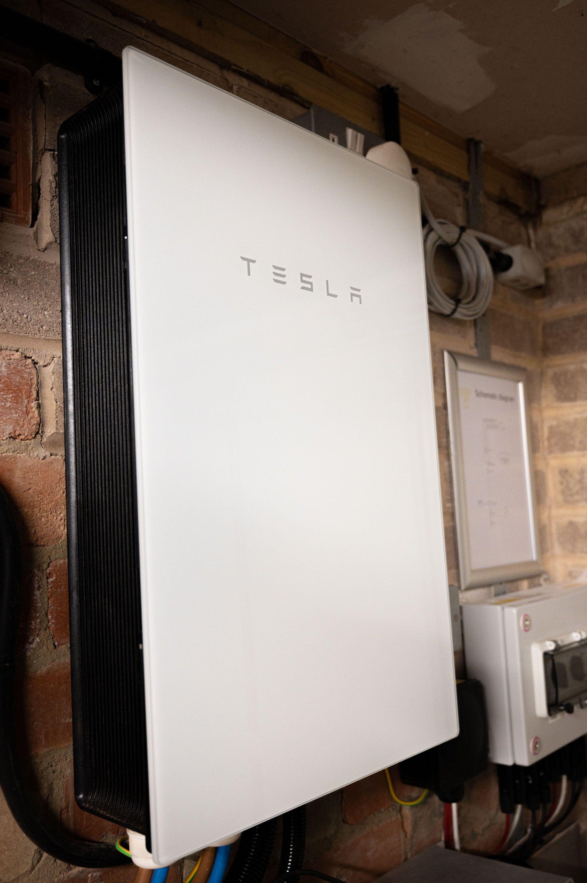 Tesla Powerwall battery mounted on a brick wall beside an isolator enclosure and a framed schematic.