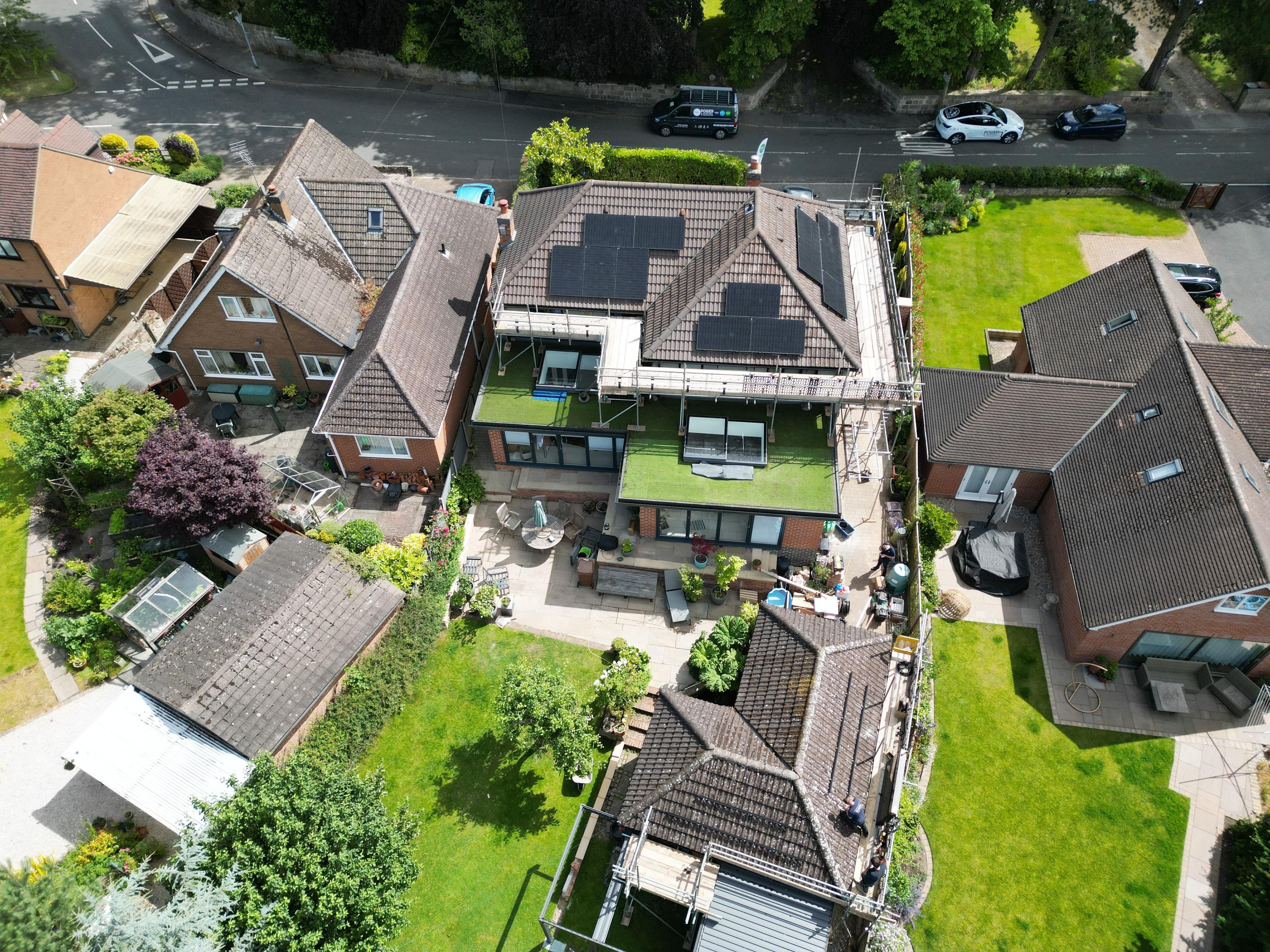 Aerial view of black solar panels on several roof faces of a house with scaffolding, among neighbouring gardens.