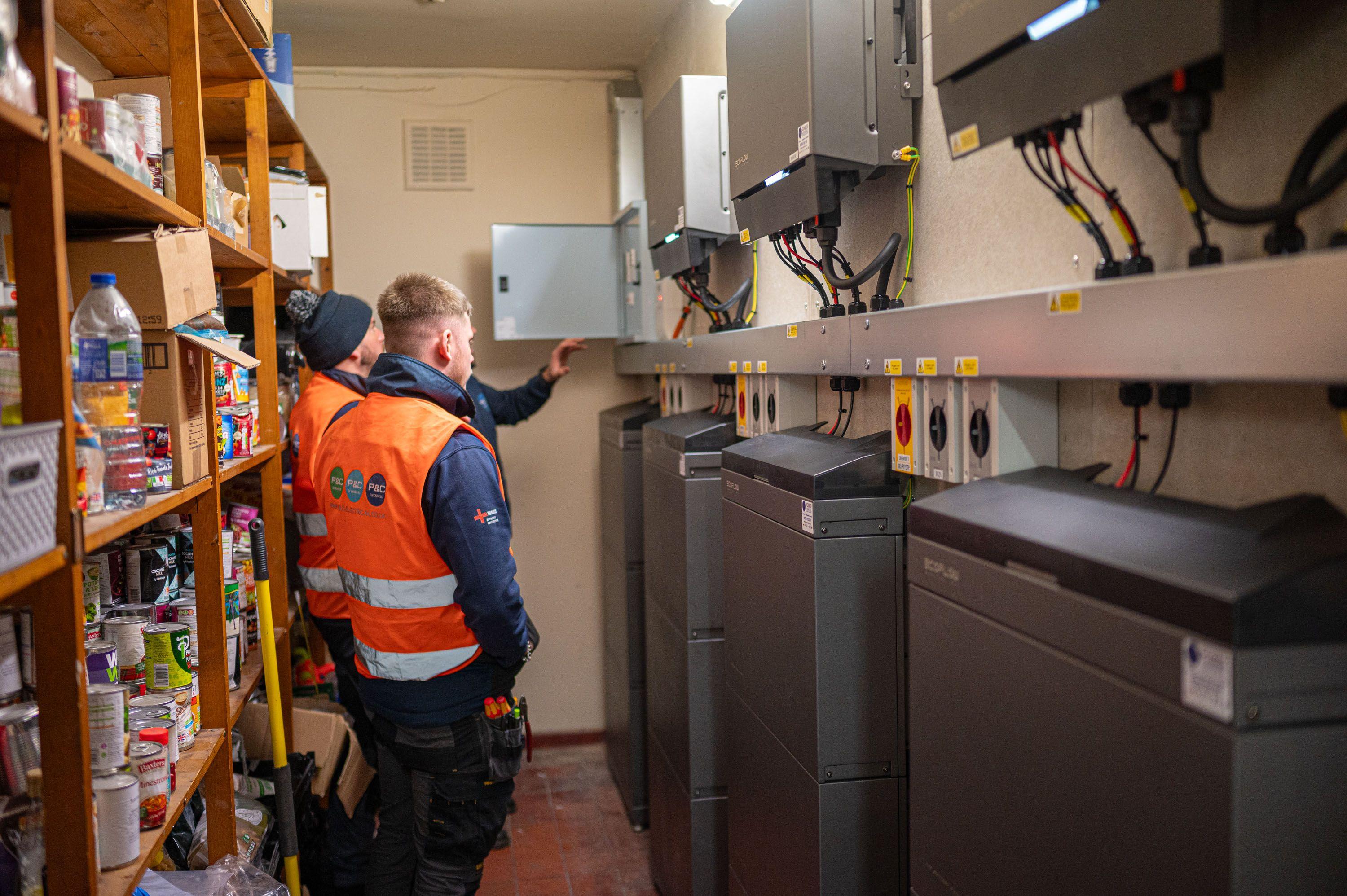 Two installers in hi-vis checking a row of inverters and battery units in a storeroom.