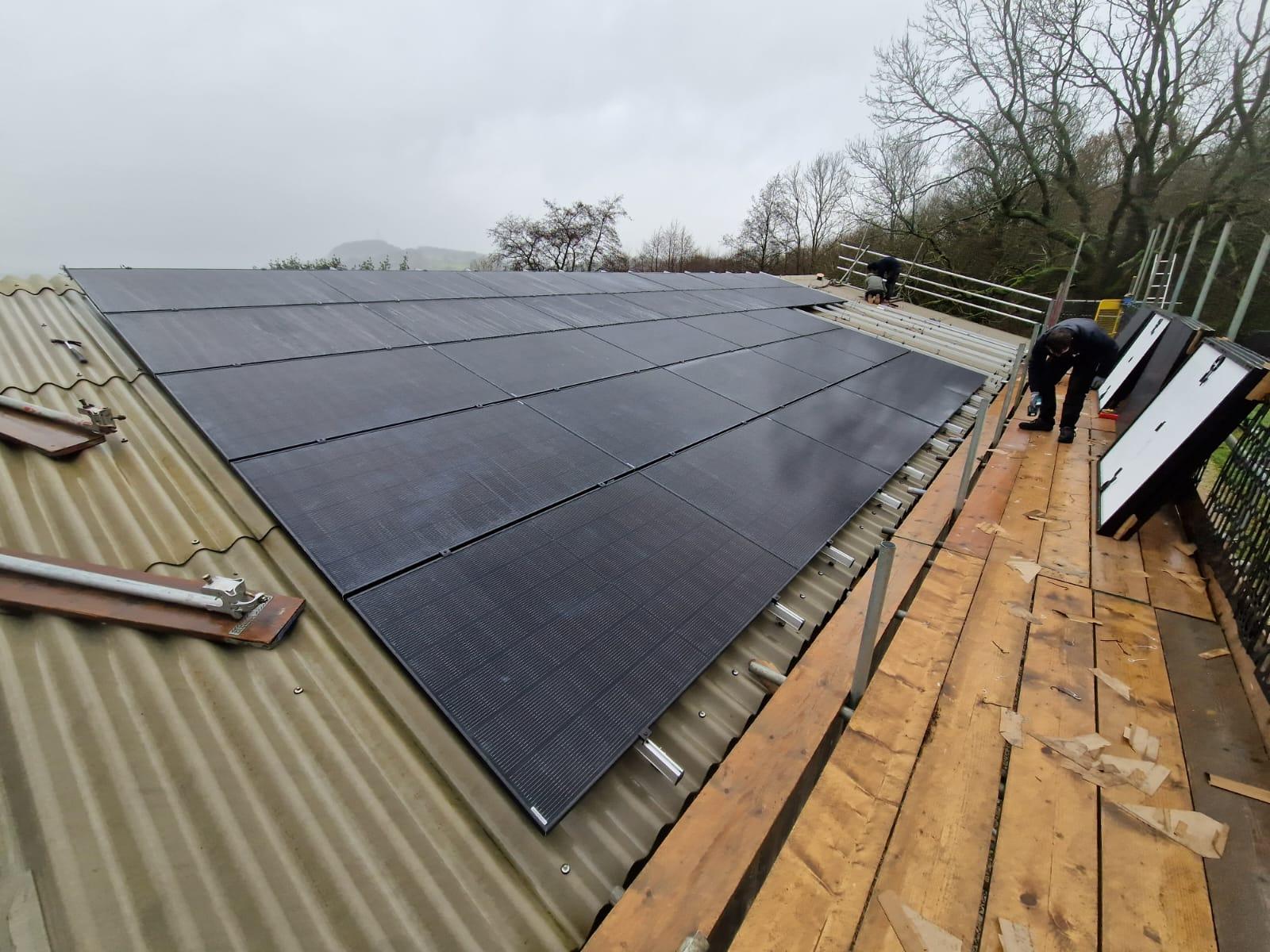 Installers working from a scaffold beside a row of black solar panels fitted to a corrugated barn roof.