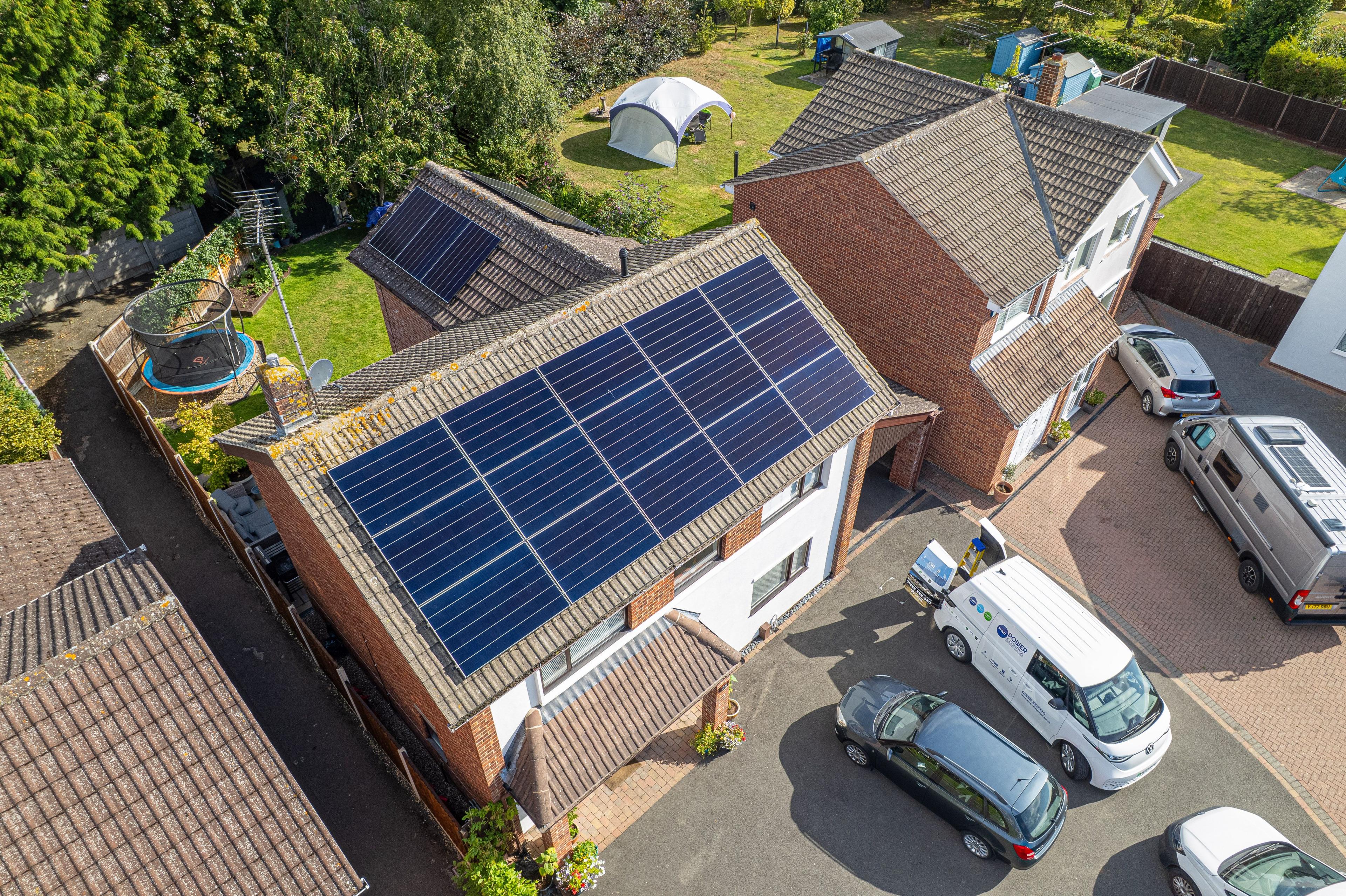 Aerial view of blue solar panels on a detached house roof, with a company van parked on the drive.