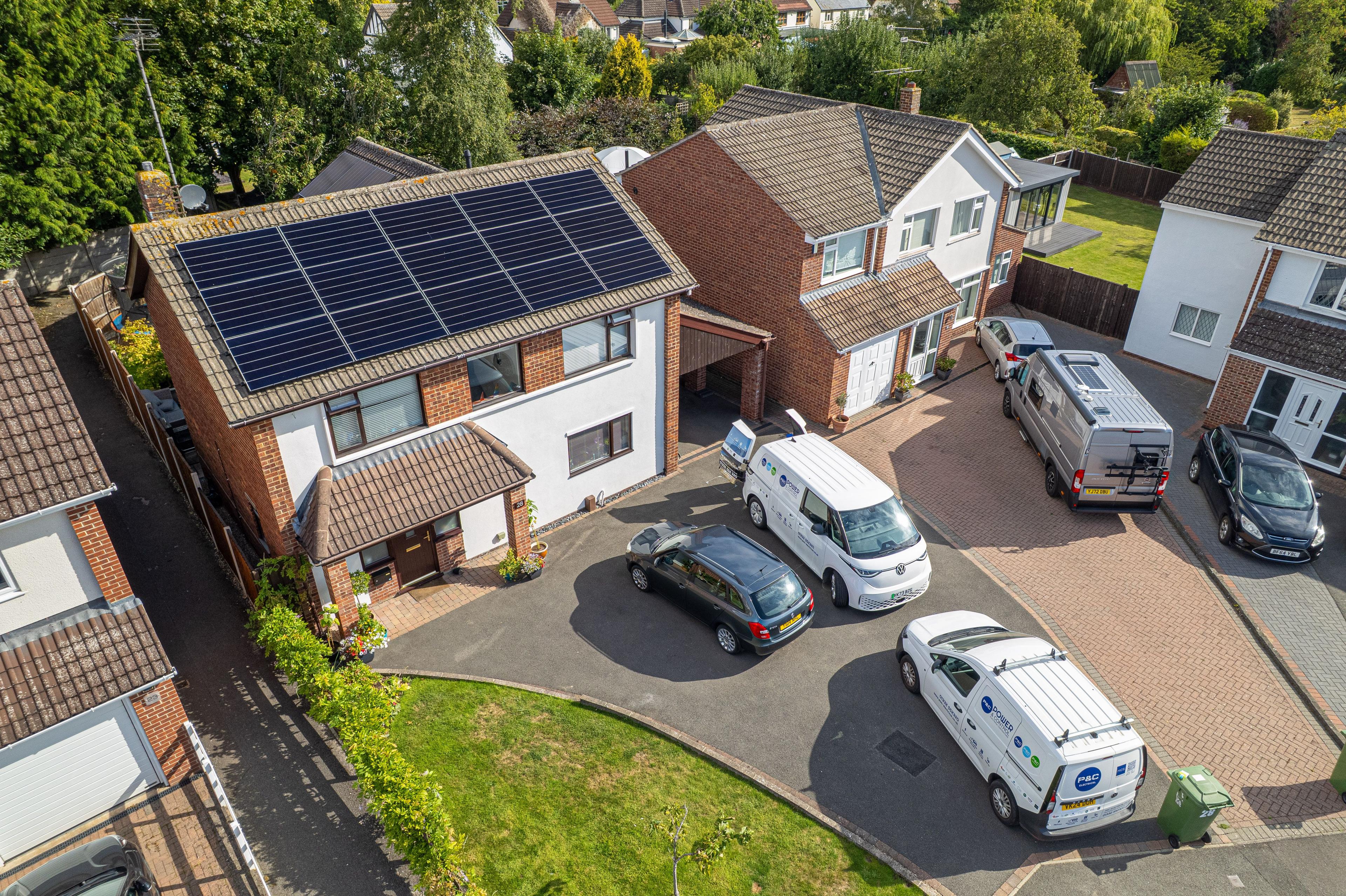 Aerial view of blue solar panels on the roof of a detached house with vans and cars on the drive.