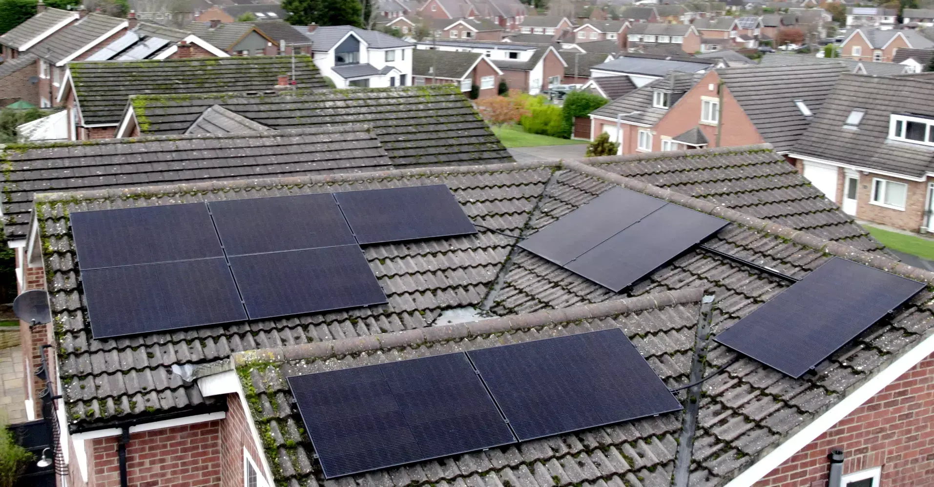 Aerial view of black solar panels on several tiled roof slopes of a house in a residential street.