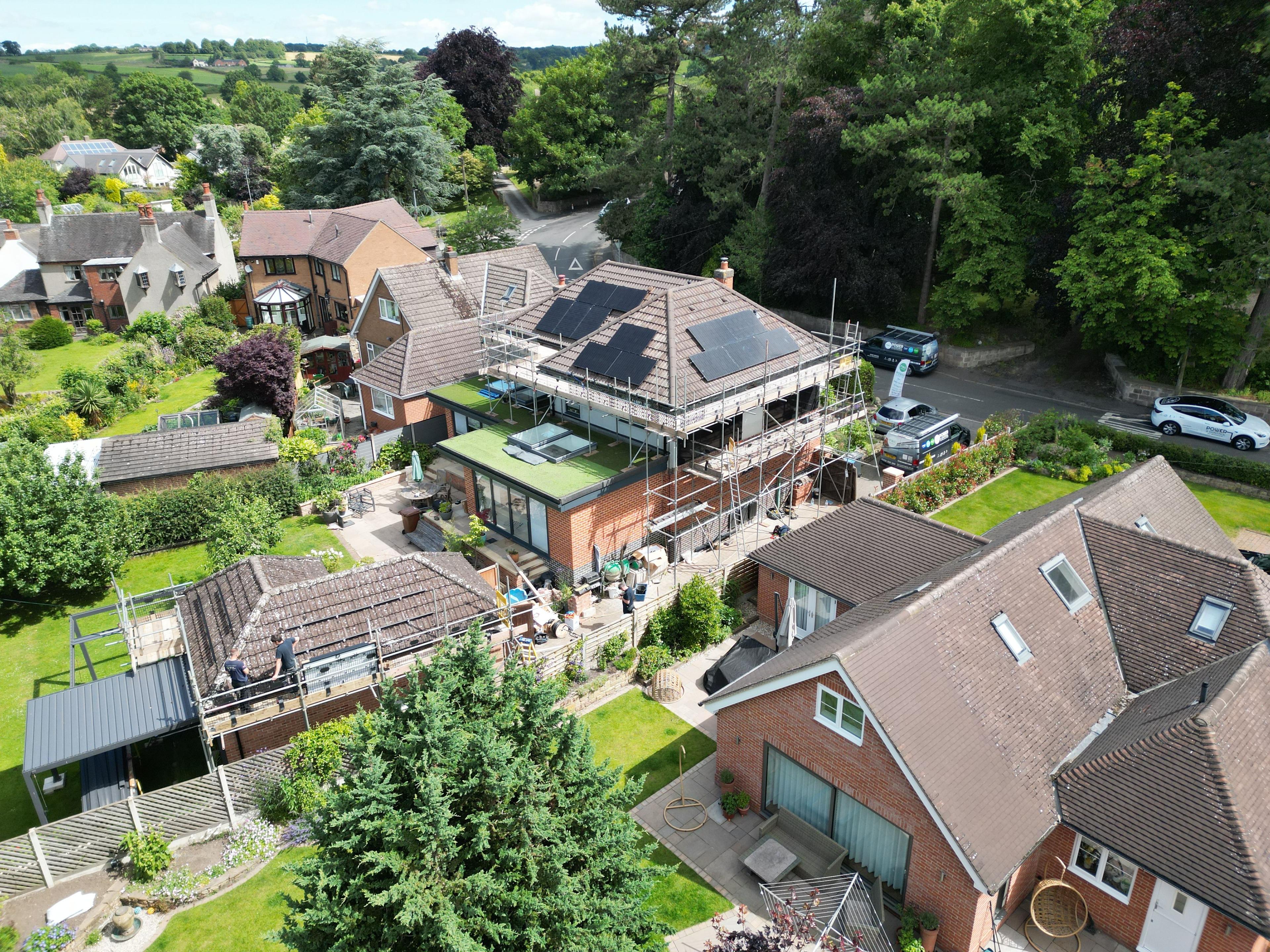Aerial view of a scaffolded house with solar panels on several roof slopes among trees and gardens.