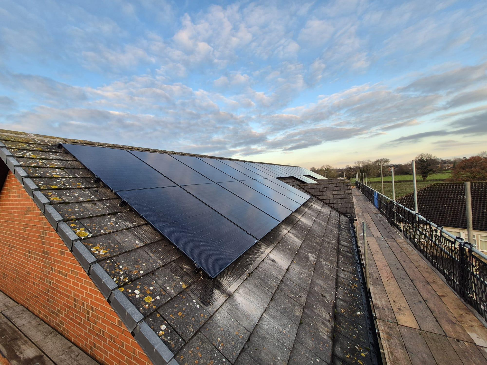 Row of black solar panels on a long tiled roof, with a scaffold walkway running alongside.