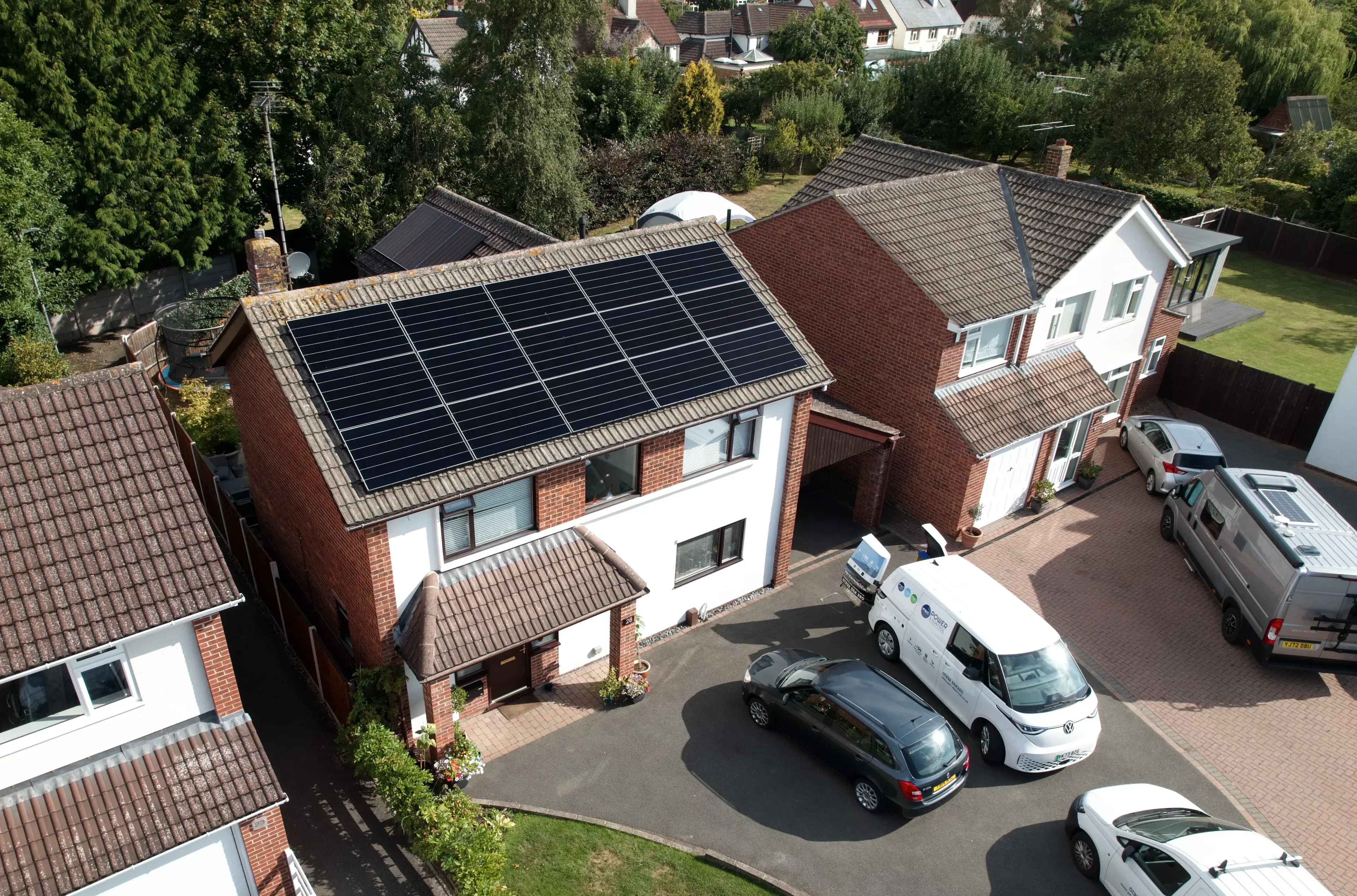 Aerial view of solar panels on a detached house roof, with a company van and cars on the drive.
