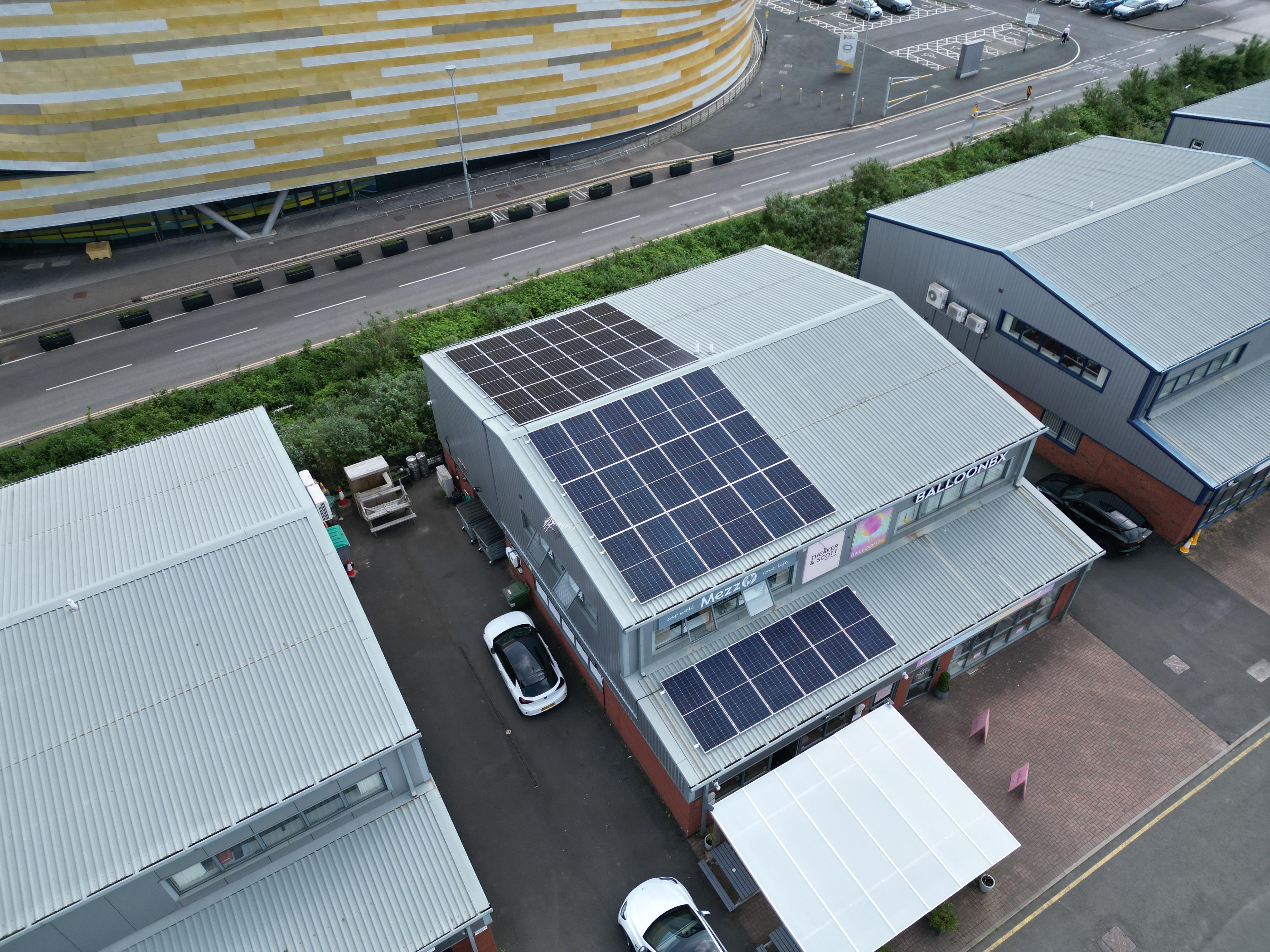 Aerial view of solar panels on a commercial unit roof, beside a main road and arena.