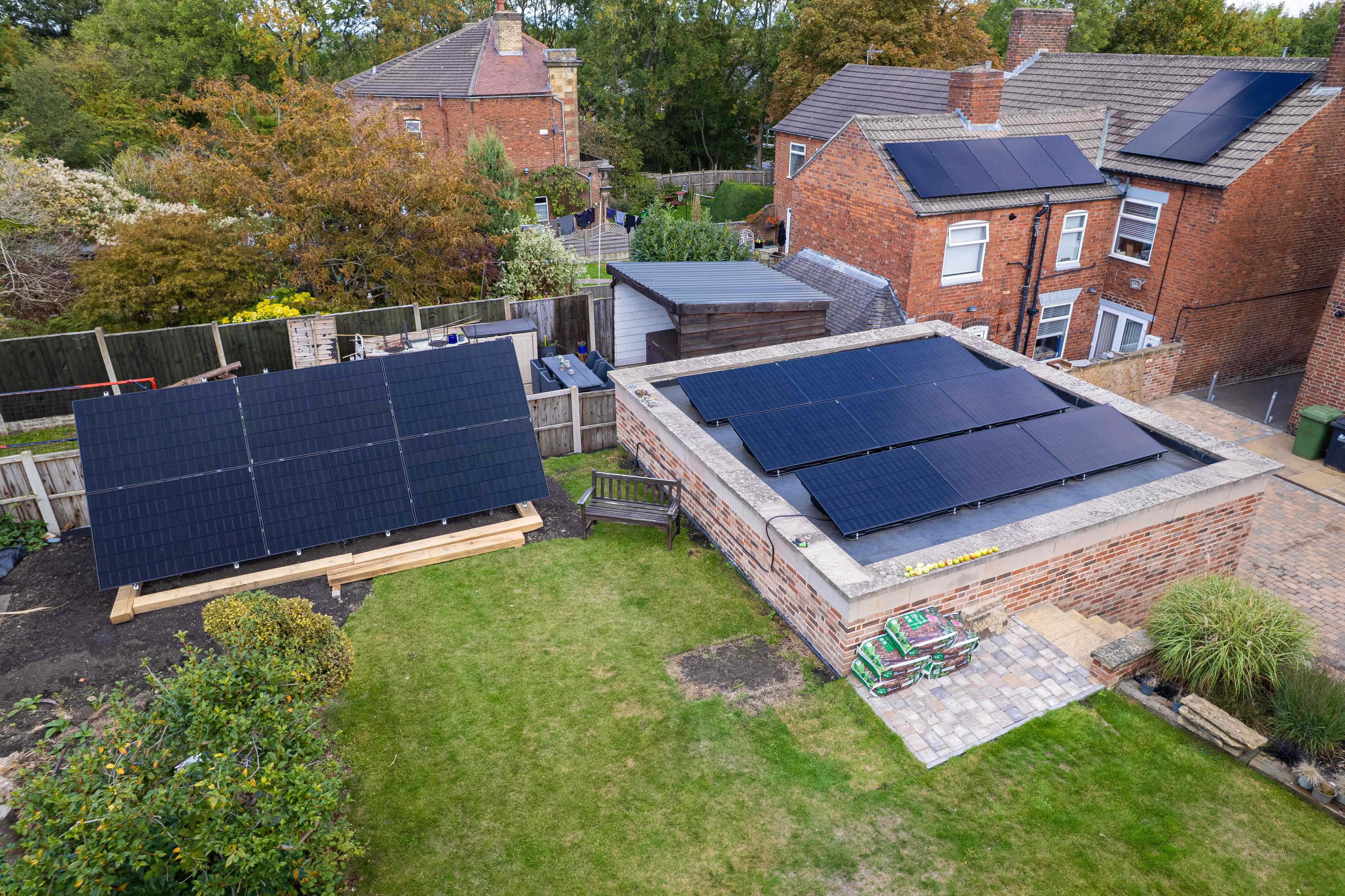 Aerial view of a ground-mounted solar array in a garden and black panels on a flat-roofed extension.