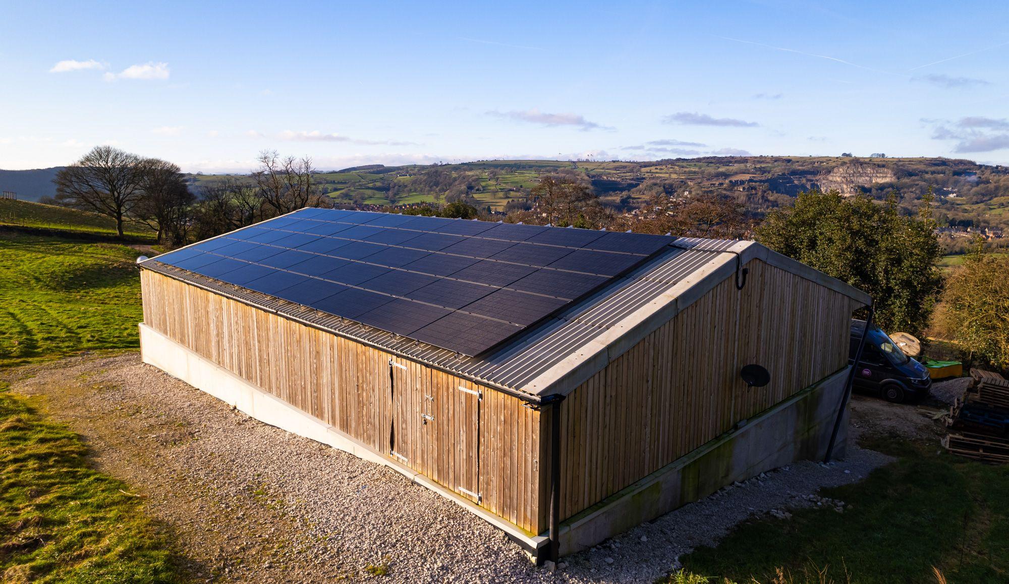 Black solar panels on the roof of a timber-clad barn overlooking rolling countryside.