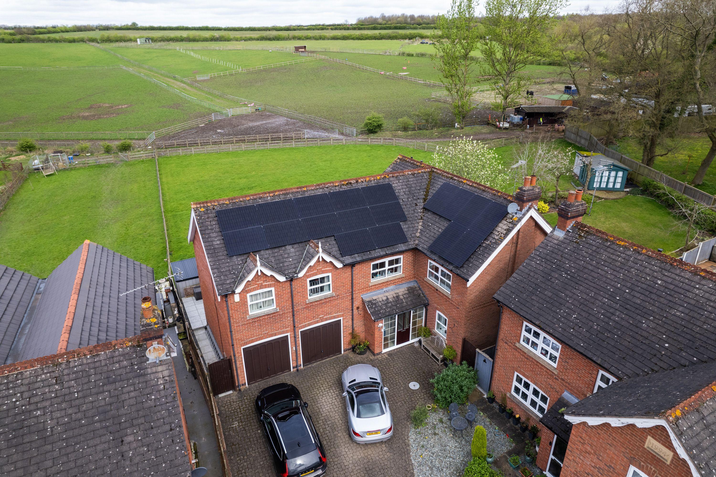Aerial view of black solar panels on two roof slopes of a red-brick detached house beside open fields.