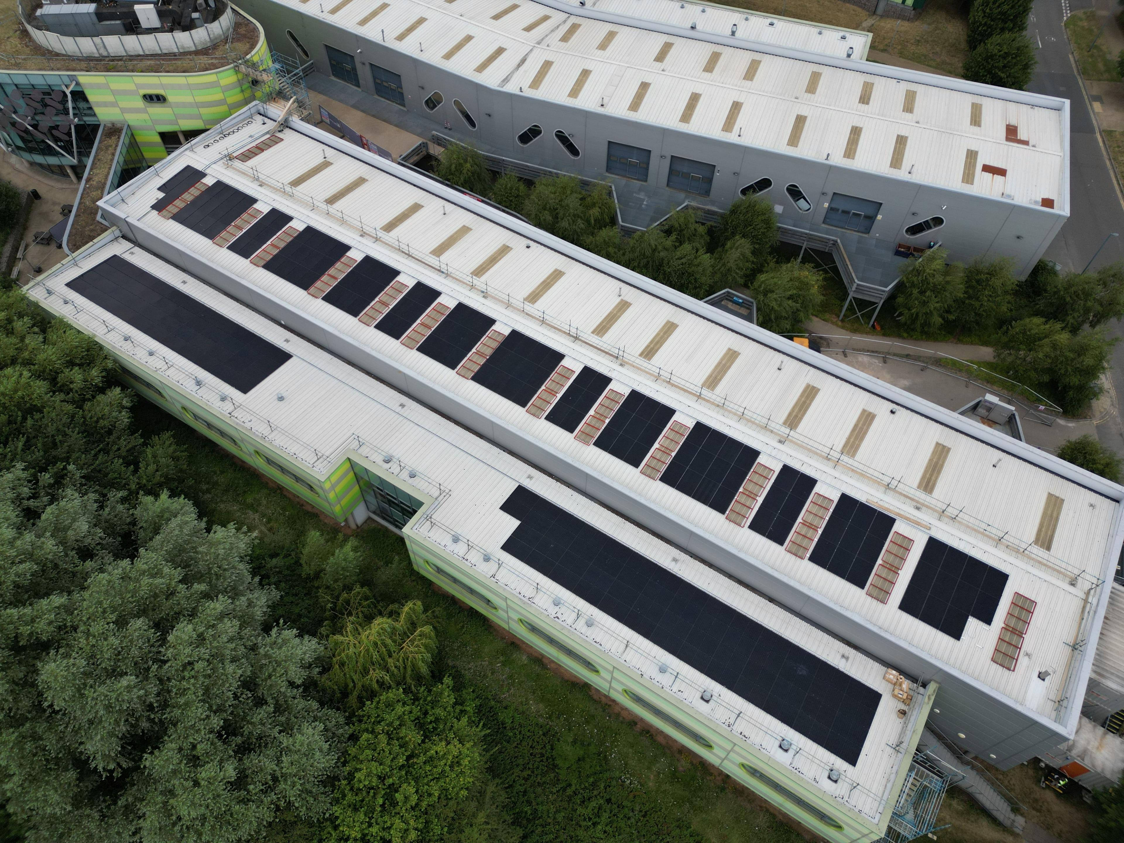 Overhead view of black solar panels fitted between rooflights on two long commercial building roofs.