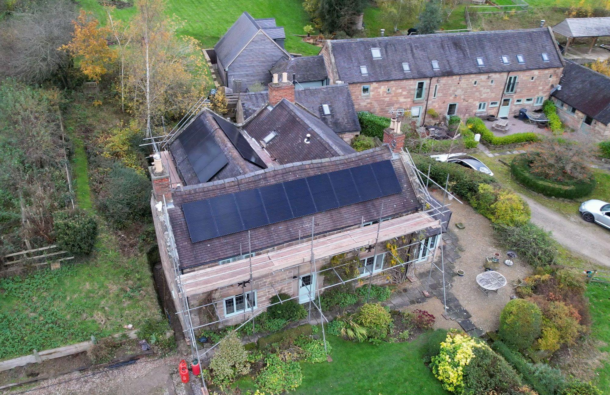 Aerial view of a stone house under scaffolding with black solar panels on two roof slopes, set among gardens.