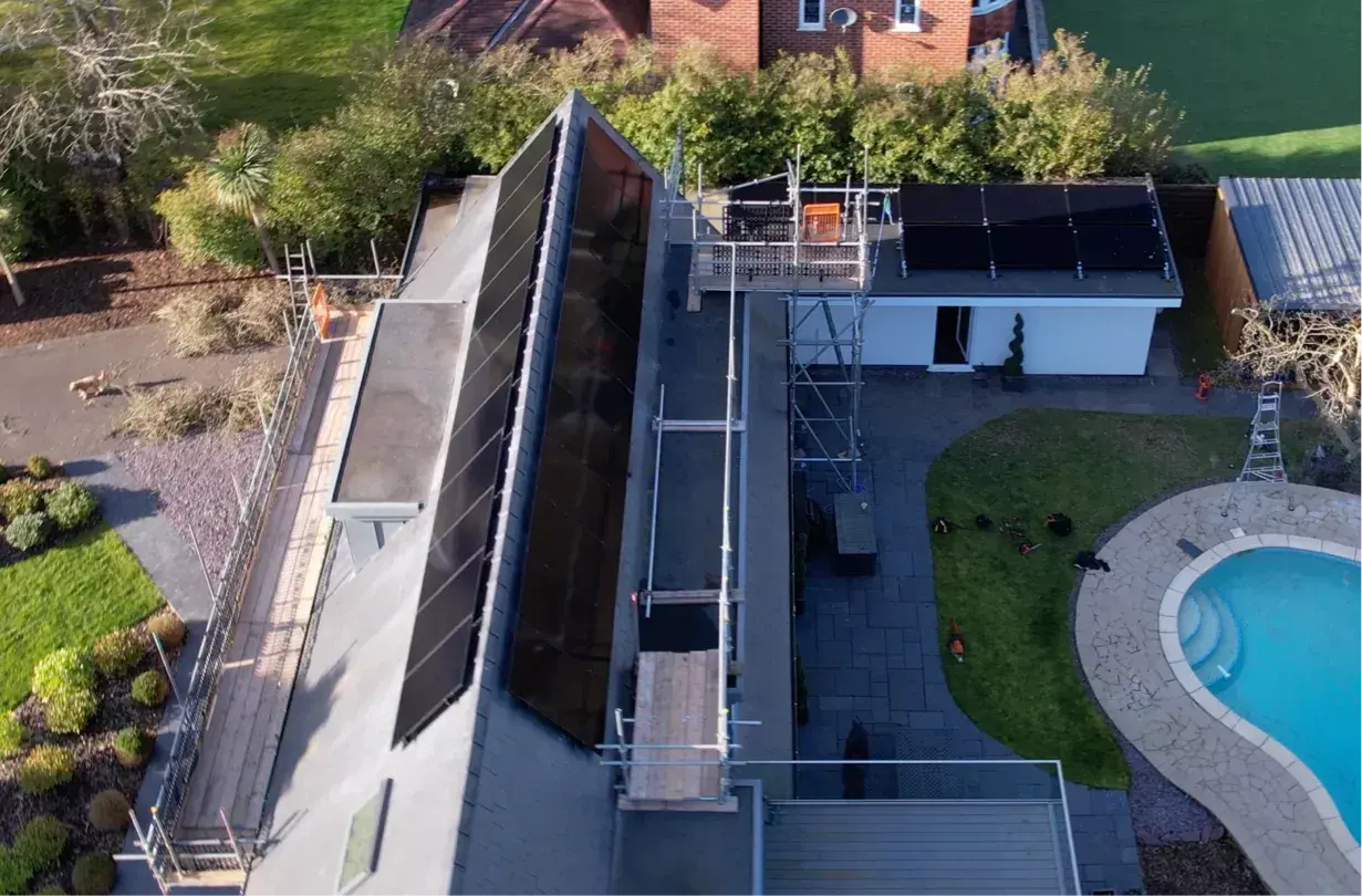 Aerial view of black solar panels on a long pitched roof and a garden outbuilding, with scaffolding beside a pool.