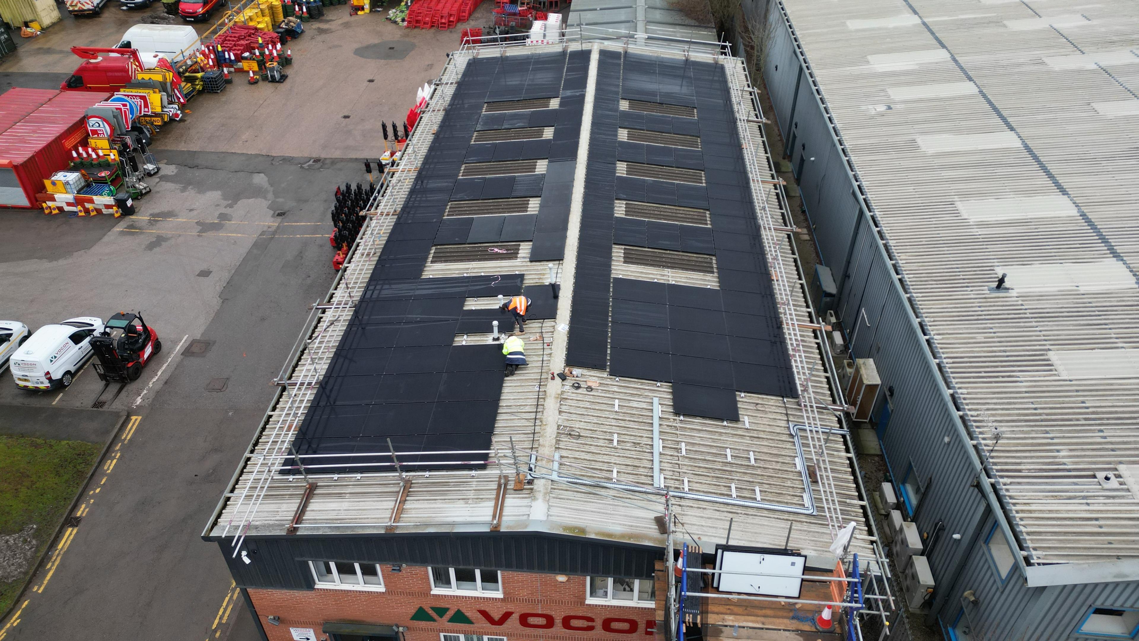 Aerial view of installers fitting black solar panels on an industrial unit roof with edge protection.