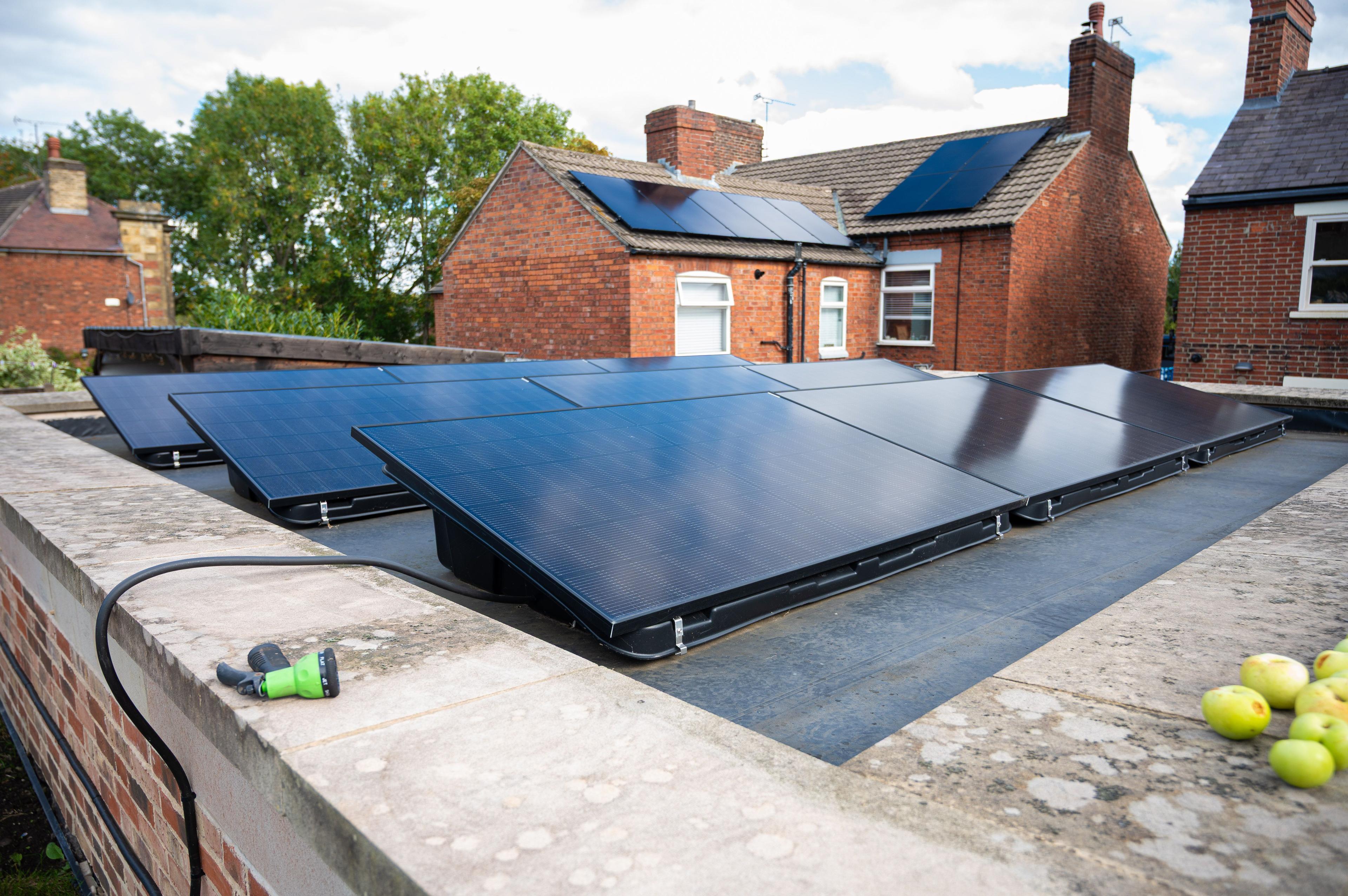 Black solar panels on low tilted mounts on a flat roof, with red-brick houses behind.