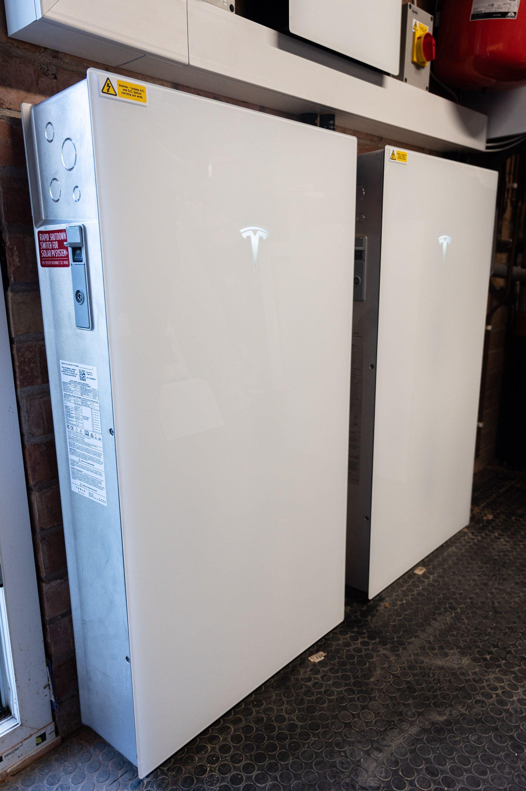 Two Tesla Powerwall batteries mounted side by side against a brick wall below an isolator and trunking.