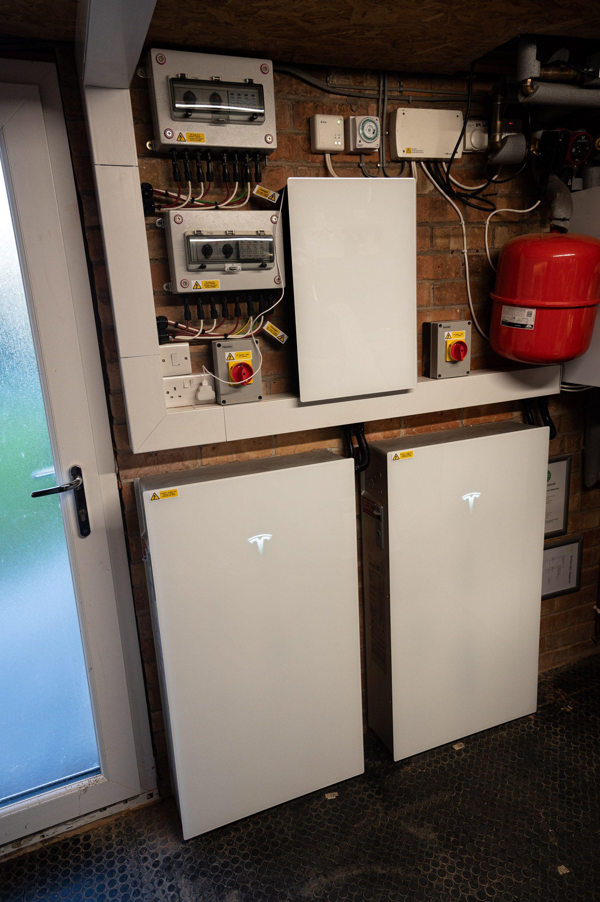 Two Tesla Powerwall batteries below a wall-mounted inverter, isolators and enclosures in a utility room.