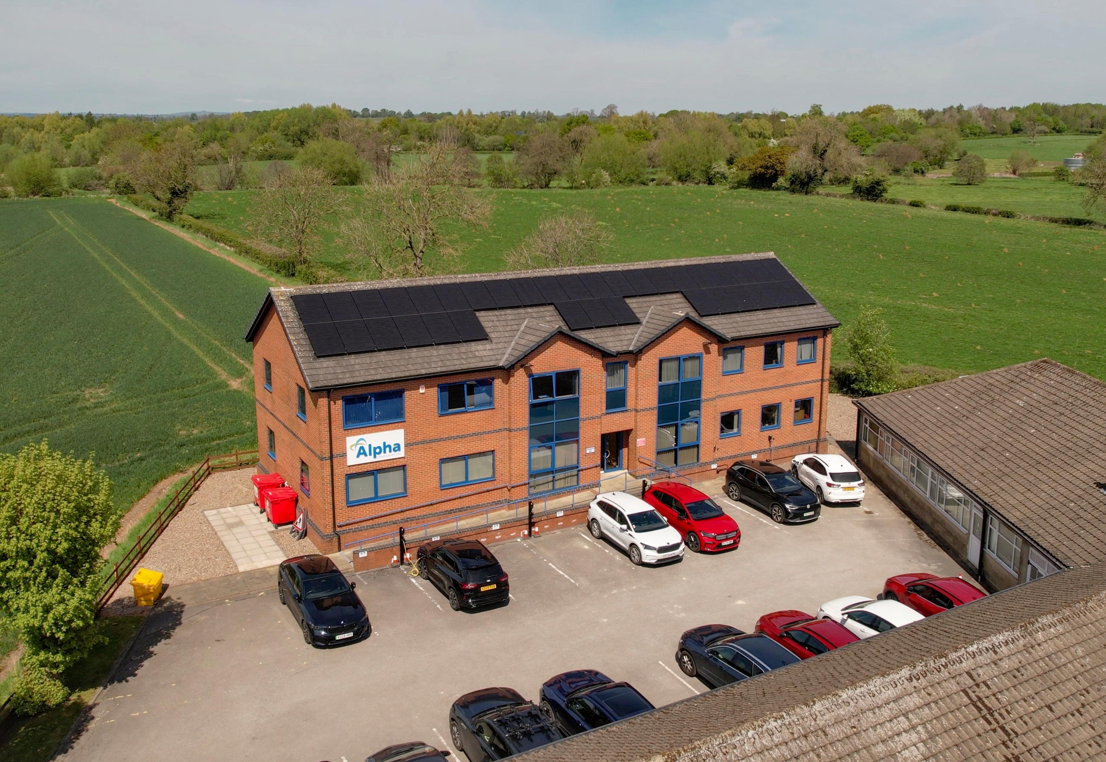 Aerial view of a brick office building with all-black solar panels on its roof beside open fields.