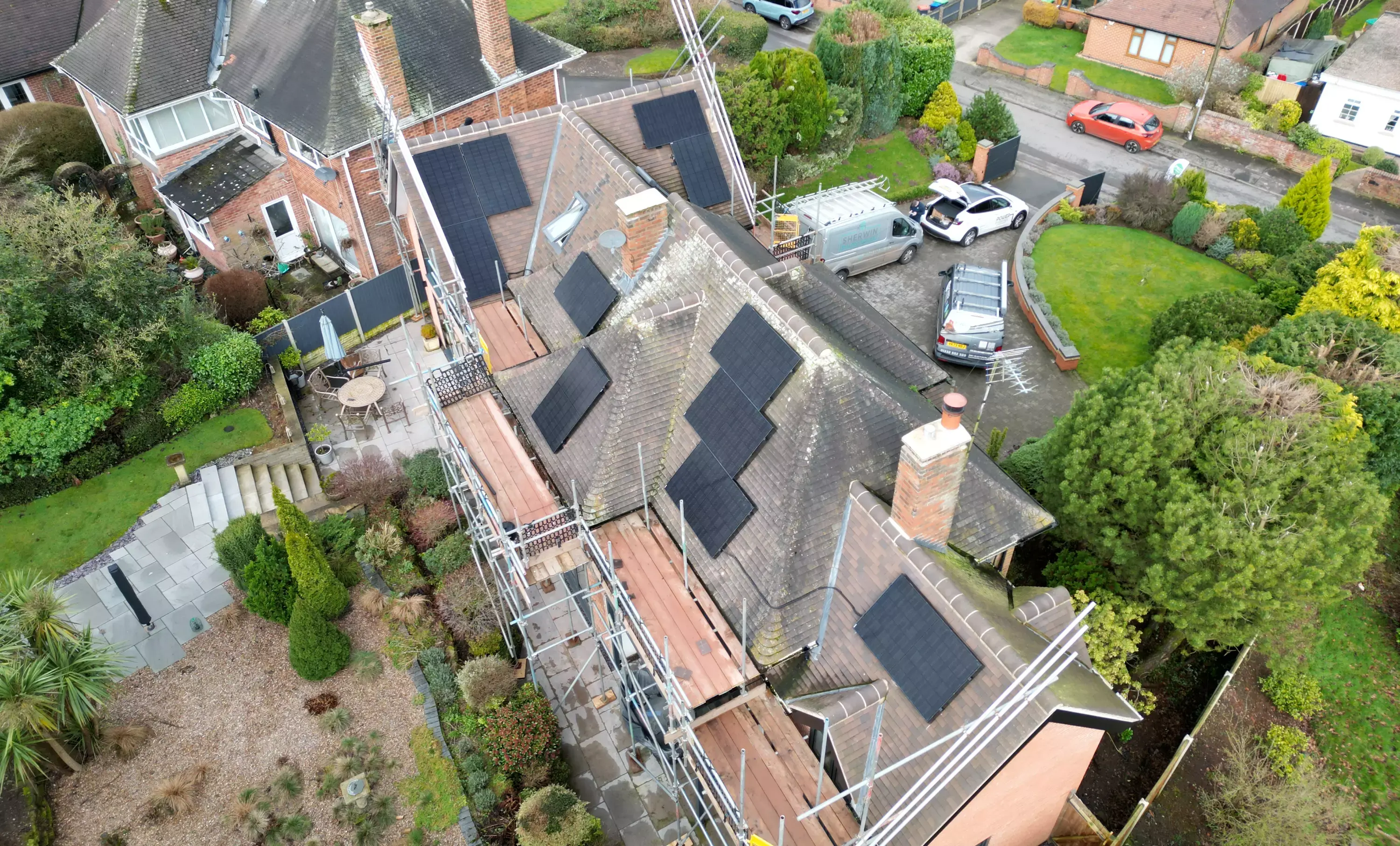 Aerial view of black solar panels spread across several roof faces of a large house wrapped in scaffolding.