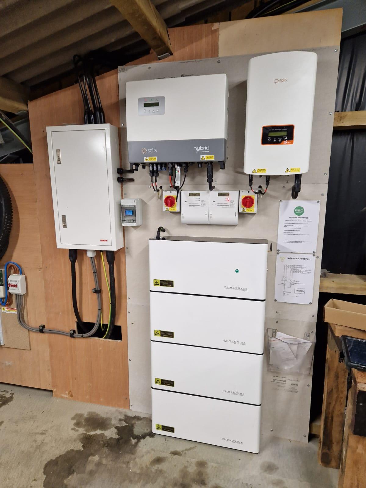Solis hybrid and solar inverters above a four-module battery stack, beside a distribution board in an outbuilding.