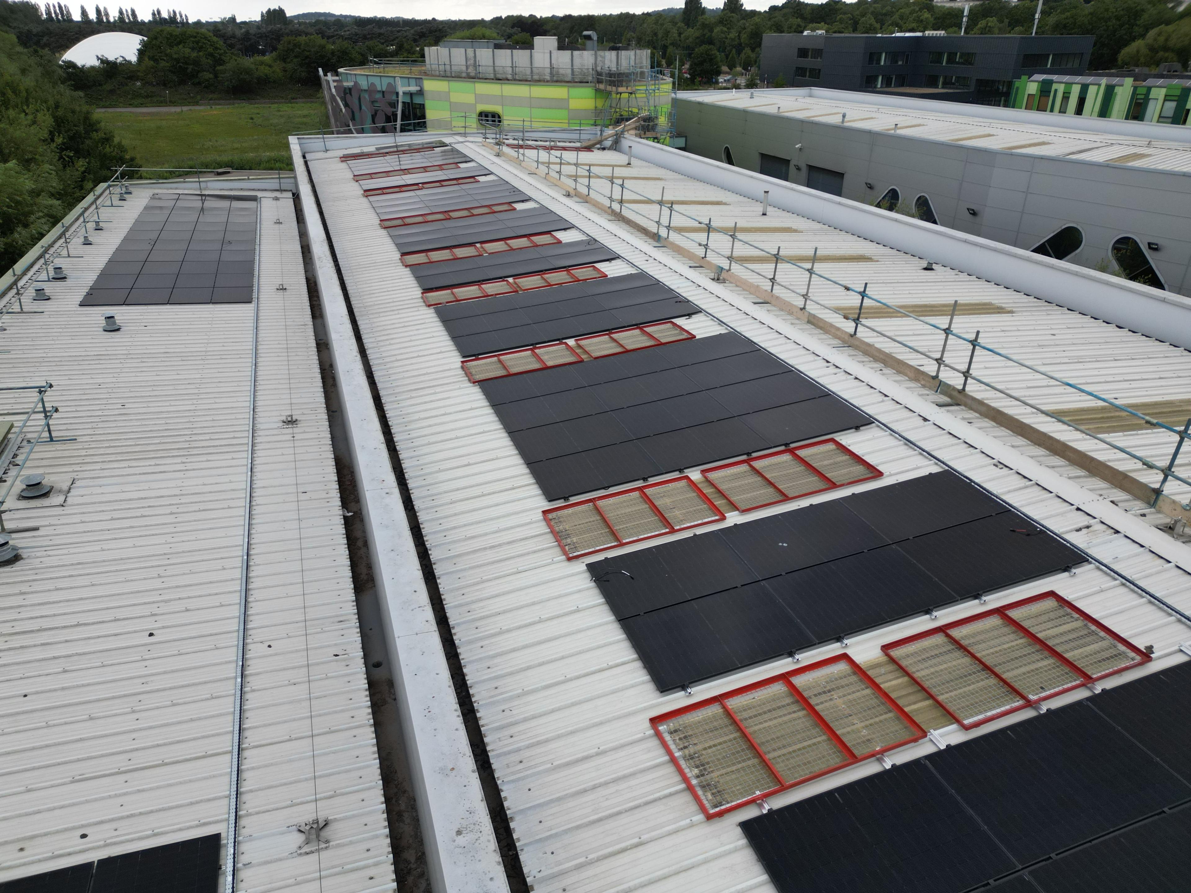 Rows of black solar panels between protected rooflights on a large commercial metal roof with edge protection.