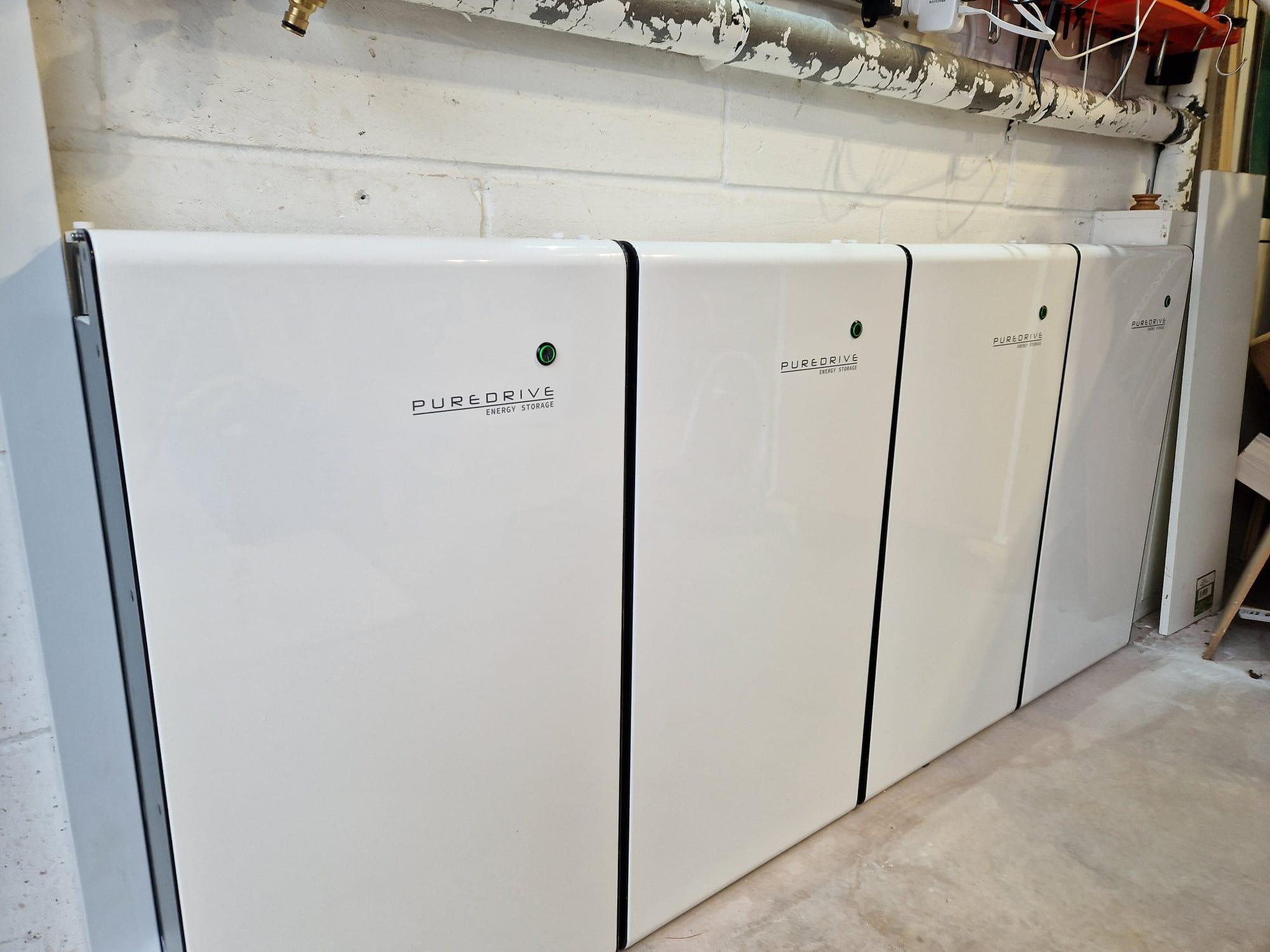 Four white PureDrive battery modules lined up along a garage wall beneath copper pipework.