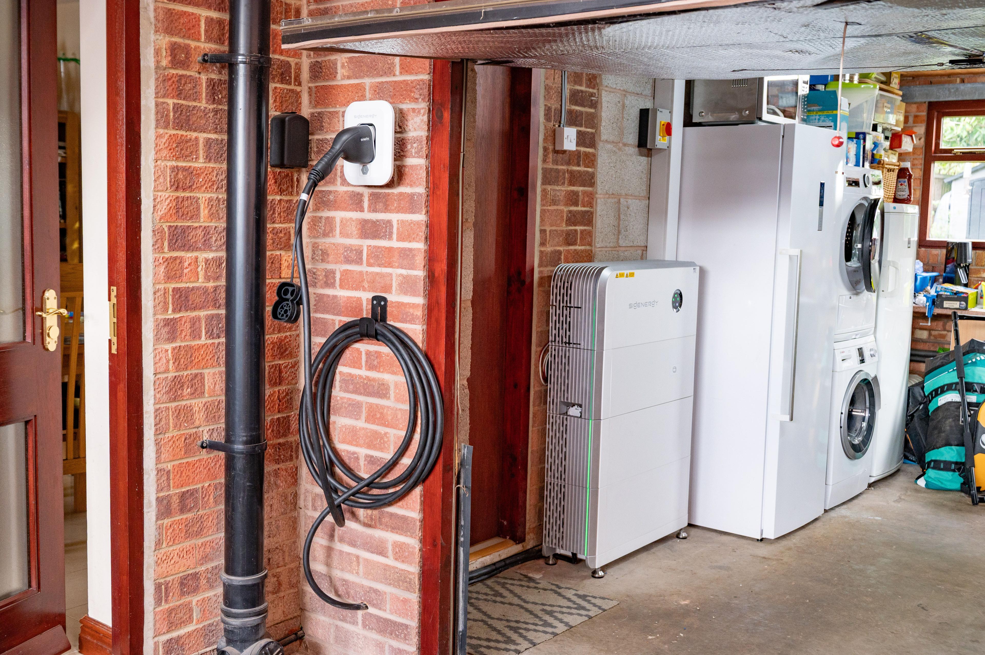 Wall-mounted EV charger with a coiled cable next to a home battery unit in a garage.
